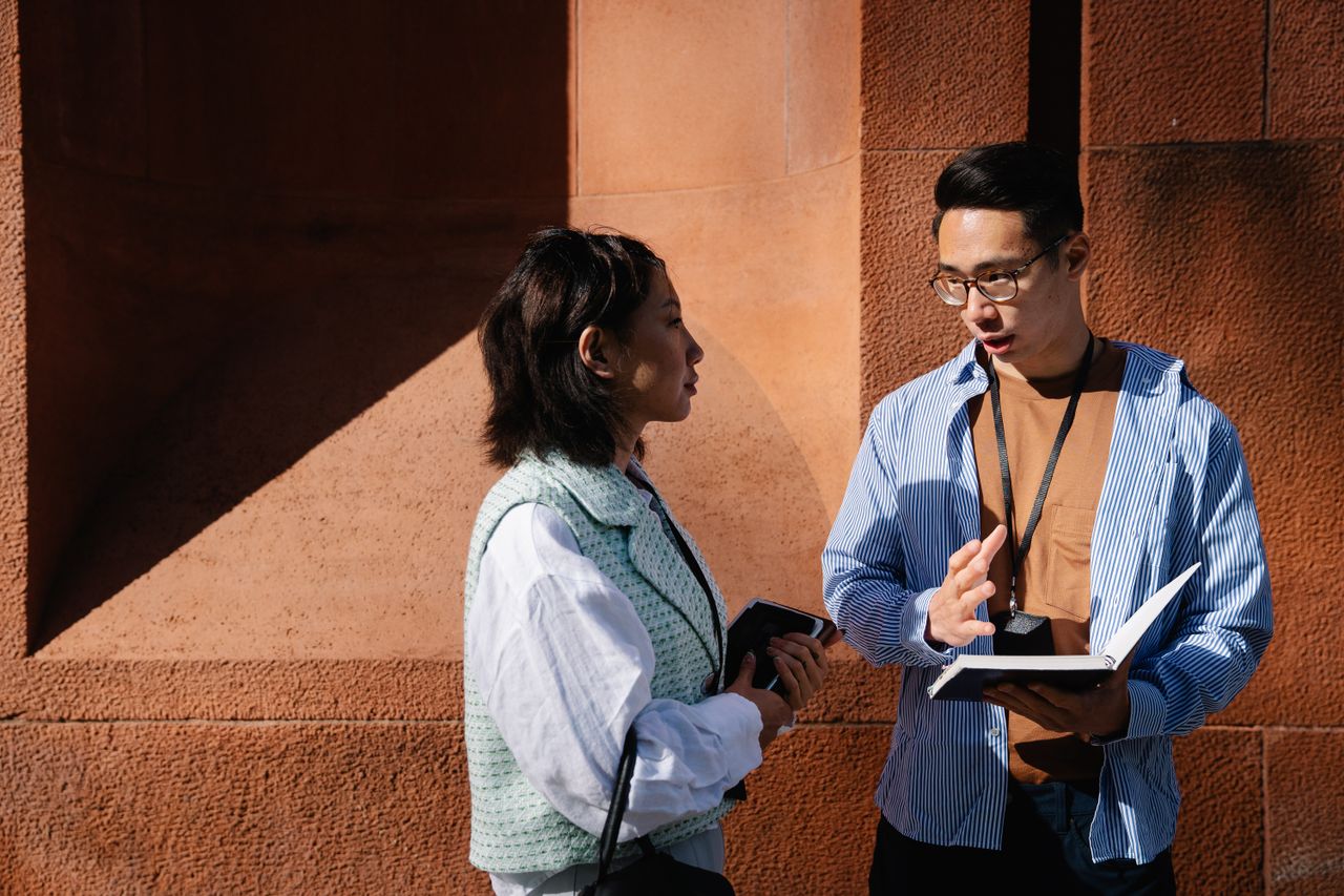 A man and woman having a conversation about work, holding notebooks in bright sunlight near a brick wall.